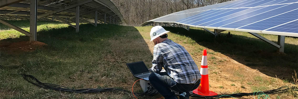 Integrator crowched down between two solar panel arrays looking at laptop screen next to orange safety cone